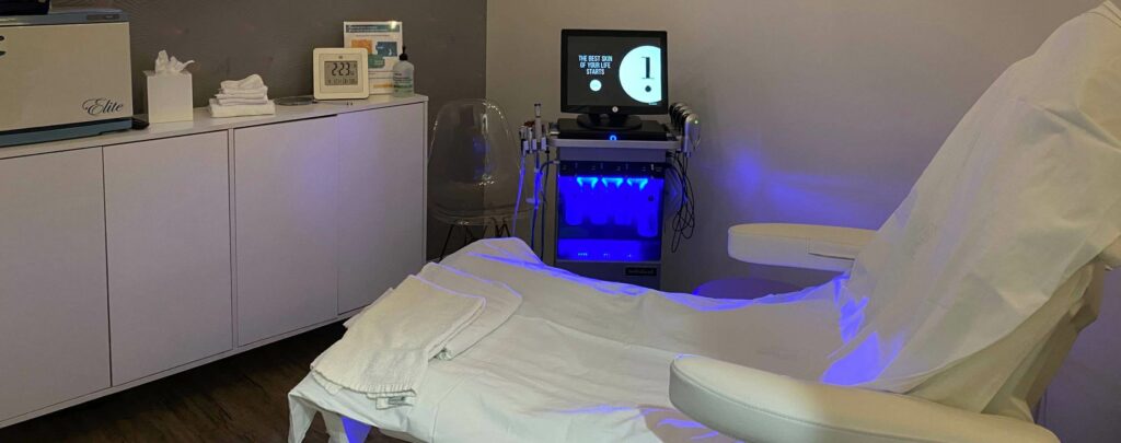 Modern aesthetic treatment room with a covered reclining chair and skincare equipment illuminated by blue LED light