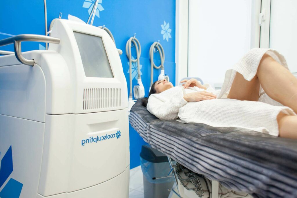 A woman undergoing a CoolSculpting session in a clinical setting, lying beside a branded fat-freezing machine