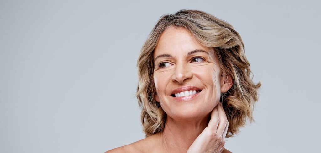 Smiling mature woman with short blonde hair gently touching her neck, showcasing healthy, radiant skin against a light background.