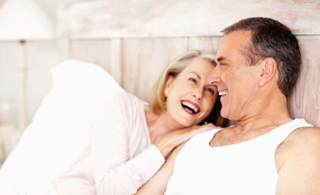 Older couple lying in bed smiling and laughing together in a bright, cozy bedroom