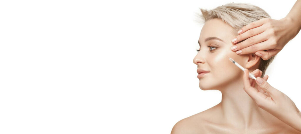 Close-up of a blonde woman receiving a cosmetic injection in her cheek while another hand gently lifts her skin