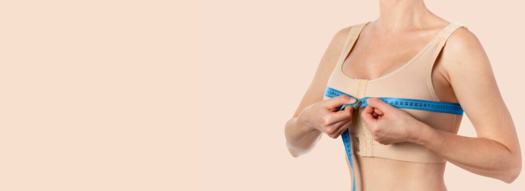 A woman in a beige bra using a blue measuring tape to measure her bust