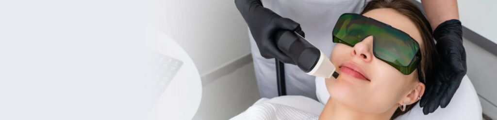 Woman receiving a laser facial treatment while wearing protective green goggles, performed by a technician in black gloves