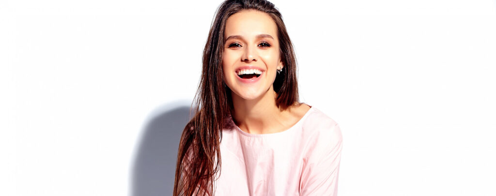 Young woman with long brown hair smiling brightly in a pink top against a white background