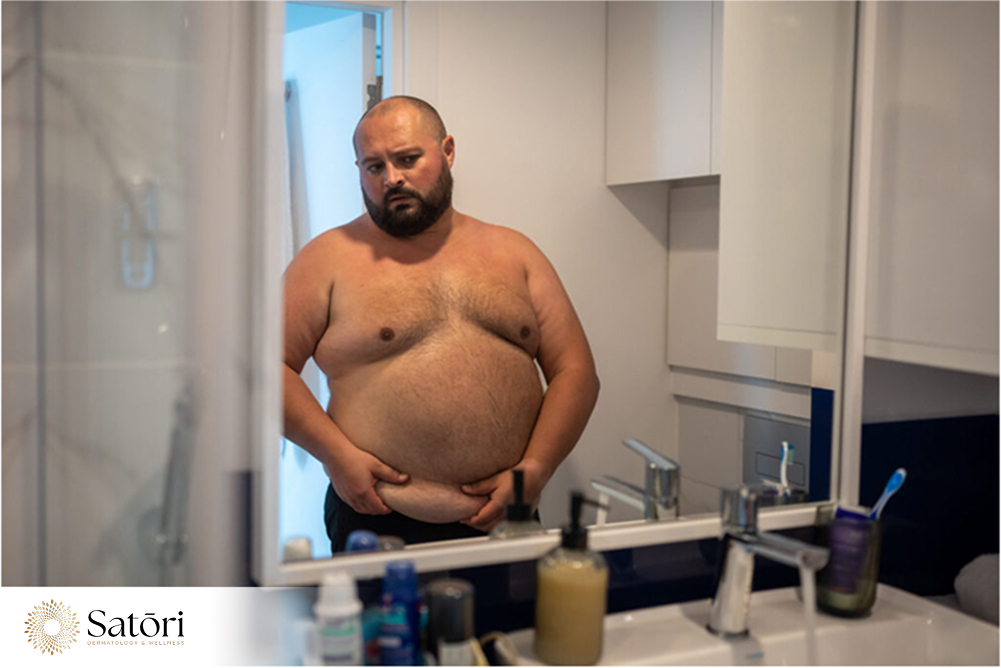 Overweight man with a large belly looking at himself in the mirror, concerned about body fat.