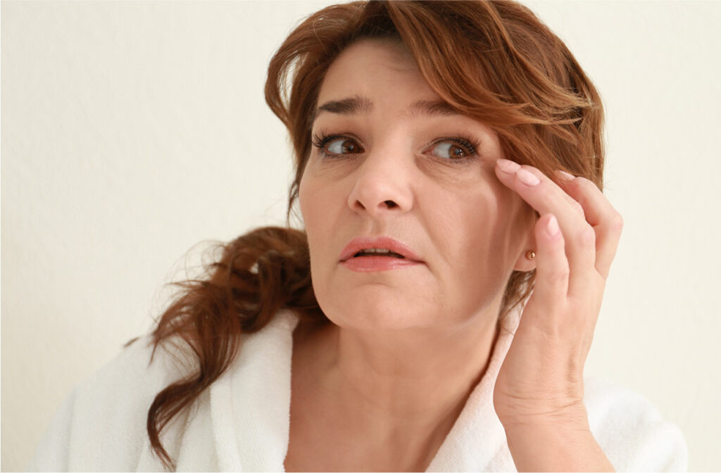 Woman gently touching the skin around her eyes while looking into a mirror, examining eye wrinkles and signs of aging.