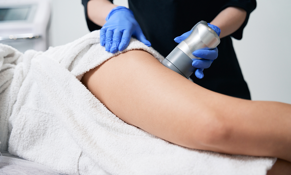 Aesthetician performing a cellulite treatment on a woman's thigh using a handheld device in a clinic setting.